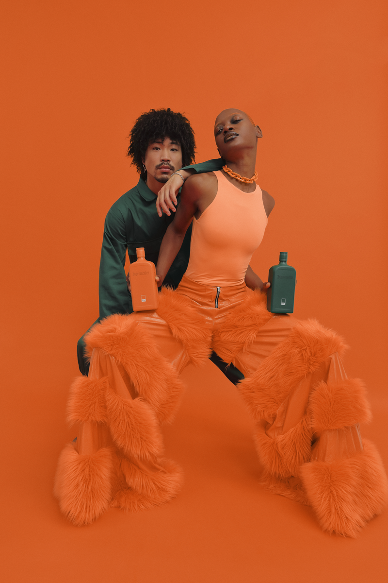 Two models pose in a studio setting with a solid orange background. One model wears a dark green top and the other wears a orange sleeveless top, a matching peach furry skirt, and accessorizes with chunky orange boots and a orange necklace.