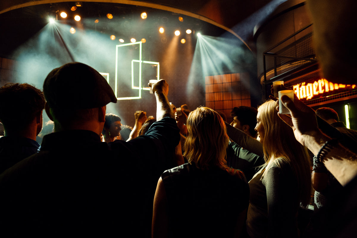 A crowd of people standing in a dimly lit nightclub or event space, facing a stage with dramatic spotlights and glowing geometric light designs. A large illuminated sign on the right appears to read Jägermeister.