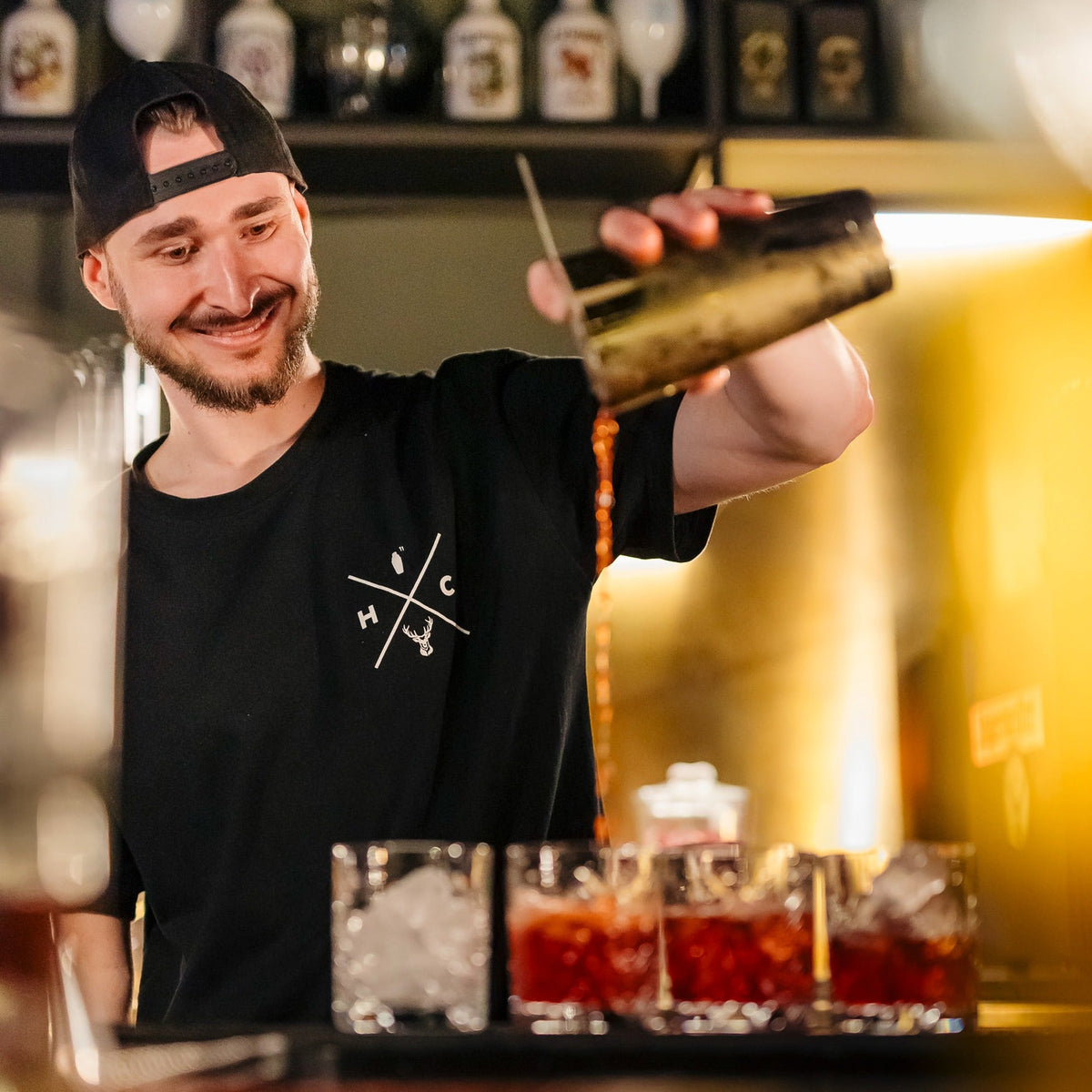 Person standing behind a bar pours a reddish cocktail from a metal shaker into multiple glasses lined up on the counter, with liquor bottles visible on shelves behind.