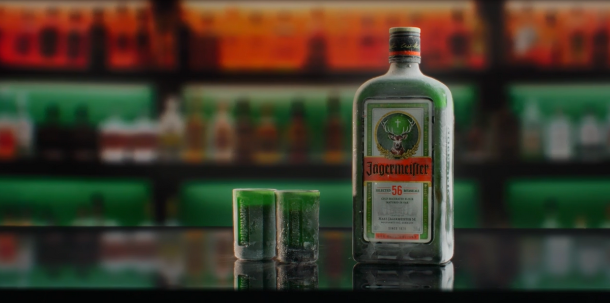 A bottle of Jägermeister stands on a reflective bar counter next to two small green shot glasses, with a blurred bar shelf background.