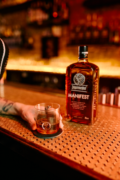 A bottle of Jagermeister Manifest and a hand holding a glass of the same amber liquor on a warm, textured bar counter in a dimly lit bar setting.