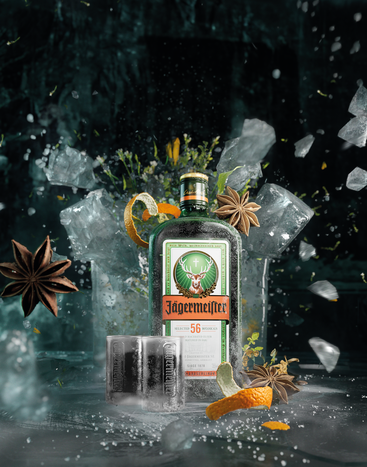 A bottle of Jägermeister bottlestands in a dramatic studio setup with crushed ice, shattered glass, star anise, orange curls, and two filled tumblers. The background is dark and moody, with floating ice fragments and a reflective surface below.