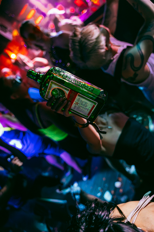 A person in a crowded nightclub or party setting holds up a Jägermeister bottle with a red and white label under colorful purple, blue, and pink party lighting.