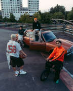 A group of four people in an urban outdoor setting with a rusted orange car, modern high-rise buildings, and a bridge in the background. One person in a bright orange sweatshirt sits on a bicycle in the foreground, another in a white Jägermeister jersey with '56' stands holding a football, and two others are positioned around the vintage car.