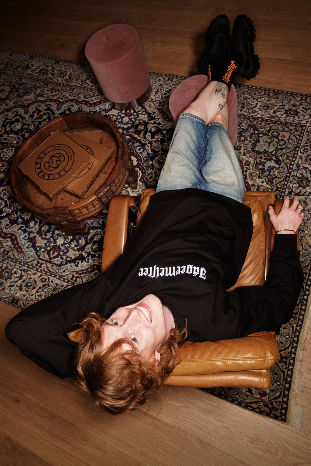 Personne allongée sur un fauteuil en cuir, les pieds posés sur un tapis, portant un sweat-shirt noir Jägermeister.