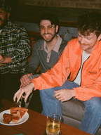 Two men sitting together, one wearing an orange Jägermeister jacket, with a plate of pizza and a glass on a table.