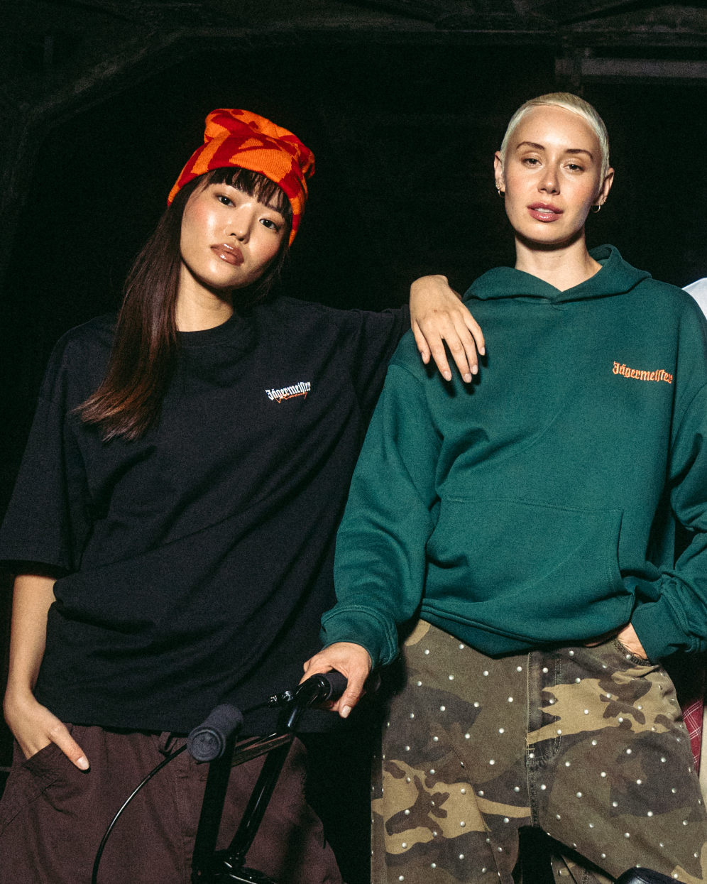 Deux jeunes femmes posent avec un vélo BMX noir devant un pont industriel sombre la nuit. Elles portent des vêtements streetwear Jägermeister, notamment un sweat à capuche vert, un T-shirt blanc, un gilet noir et un pantalon cargo.