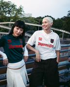 Quatre personnes se tiennent en extérieur sur un pont ou une passerelle, vêtues de streetwear. L’une porte une veste zippée orange vif, une autre porte un maillot Jägermeister vert foncé avec une jupe blanche, une troisième porte un maillot Jägermeister blanc avec un short foncé, et une quatrième porte un sweat à capuche noir et une casquette orange.