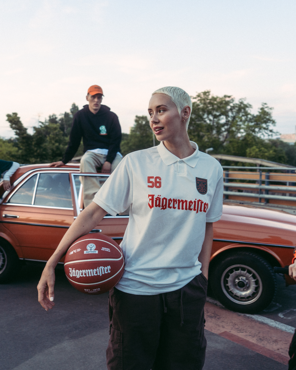 Un groupe de quatre personnes pose en extérieur sur un pont ou un passage supérieur, avec une voiture vintage orange derrière eux. La personne au centre tient un ballon de basket et porte un maillot Jägermeister blanc, tandis que les autres portent du streetwear orange vif, vert et noir.
