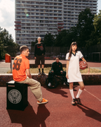 Scène lifestyle streetwear en extérieur sur un terrain de basket, avec un grand immeuble d’habitation en arrière-plan. Un homme assis en maillot Jägermeister orange est installé sur une boîte noire ressemblant à un haut-parleur, tandis qu’une femme debout en robe polo blanche Jägermeister tient un ballon de basket.