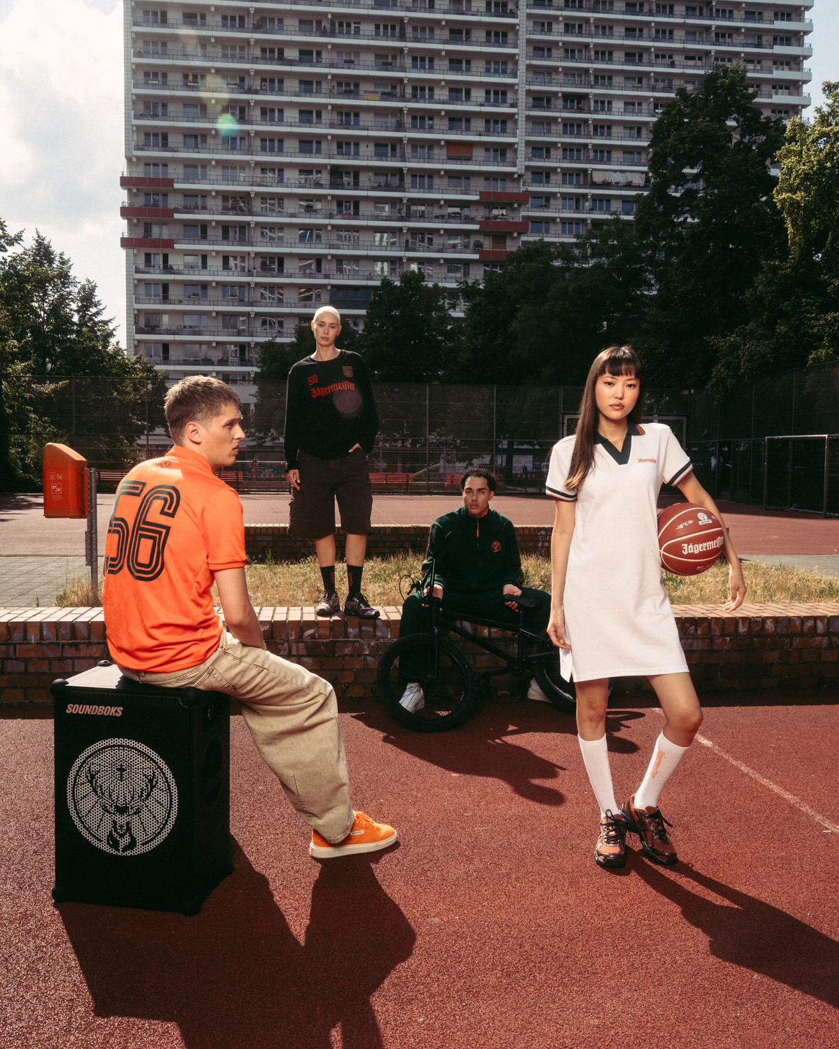 Scène lifestyle streetwear en extérieur sur un terrain de basket, avec un grand immeuble d’habitation en arrière-plan. Un homme assis en maillot Jägermeister orange est installé sur une boîte noire ressemblant à un haut-parleur, tandis qu’une femme debout en robe polo blanche Jägermeister tient un ballon de basket.