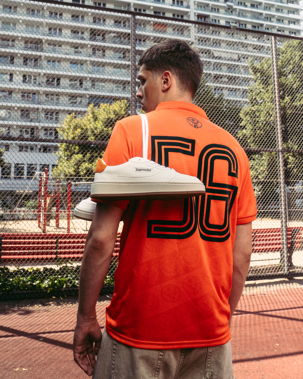Une personne se tient sur un terrain de sport extérieur en portant un maillot Jägermeister orange vif avec un grand « 56 » imprimé dans le dos. Une paire de baskets Jägermeister blanches est drapée sur une épaule, avec un grillage et des immeubles d’habitation en arrière-plan.