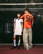 Deux mannequins se tiennent sur un court de tennis rouge devant un grillage. Le mannequin à gauche porte une robe polo de sport Jägermeister blanche avec bordures vert foncé, des chaussettes blanches et des chaussures noires.