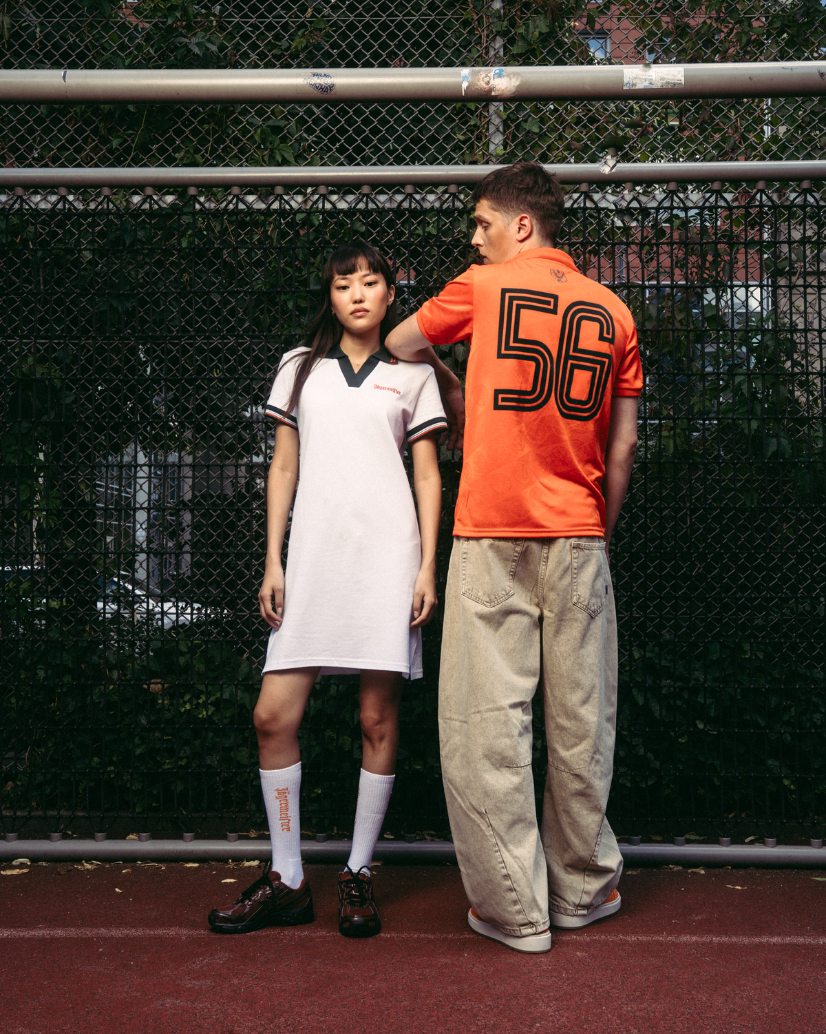 Deux mannequins se tiennent sur un court de tennis rouge devant un grillage. Le mannequin à gauche porte une robe polo de sport Jägermeister blanche avec bordures vert foncé, des chaussettes blanches et des chaussures noires.