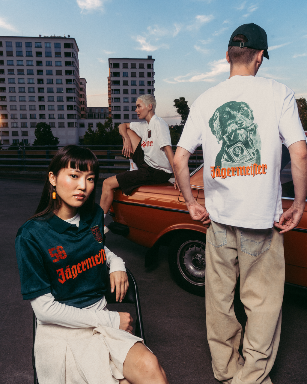 Trois mannequins portant des vêtements de marque Jägermeister dans un cadre urbain en extérieur, avec une voiture vintage orange et des immeubles d’habitation en arrière-plan. Un mannequin est assis au premier plan avec un maillot Jägermeister vert foncé, un autre est debout de dos avec un T-shirt blanc à motif graphique et un pantalon beige, et un troisième mannequin est assis sur la voiture avec un T-shirt blanc.