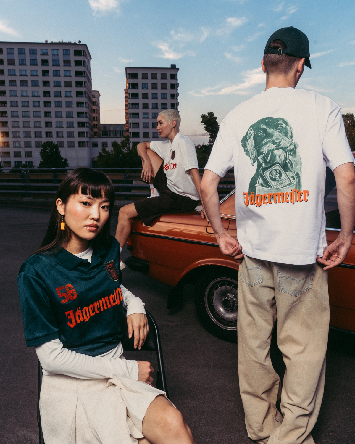 Trois mannequins portant des vêtements de marque Jägermeister dans un cadre urbain en extérieur, avec une voiture vintage orange et des immeubles d’habitation en arrière-plan. Un mannequin est assis au premier plan avec un maillot Jägermeister vert foncé, un autre est debout de dos avec un T-shirt blanc à motif graphique et un pantalon beige, et un troisième mannequin est assis sur la voiture avec un T-shirt blanc.