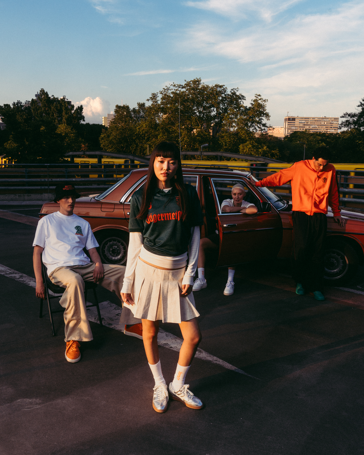 A group of four young people posing outdoors on a racetrack at sunset next to a vintage red car. The central figure is wearing a dark green Jäermeister jersey with orange lettering, a pleated white skirt, and white sneakers.