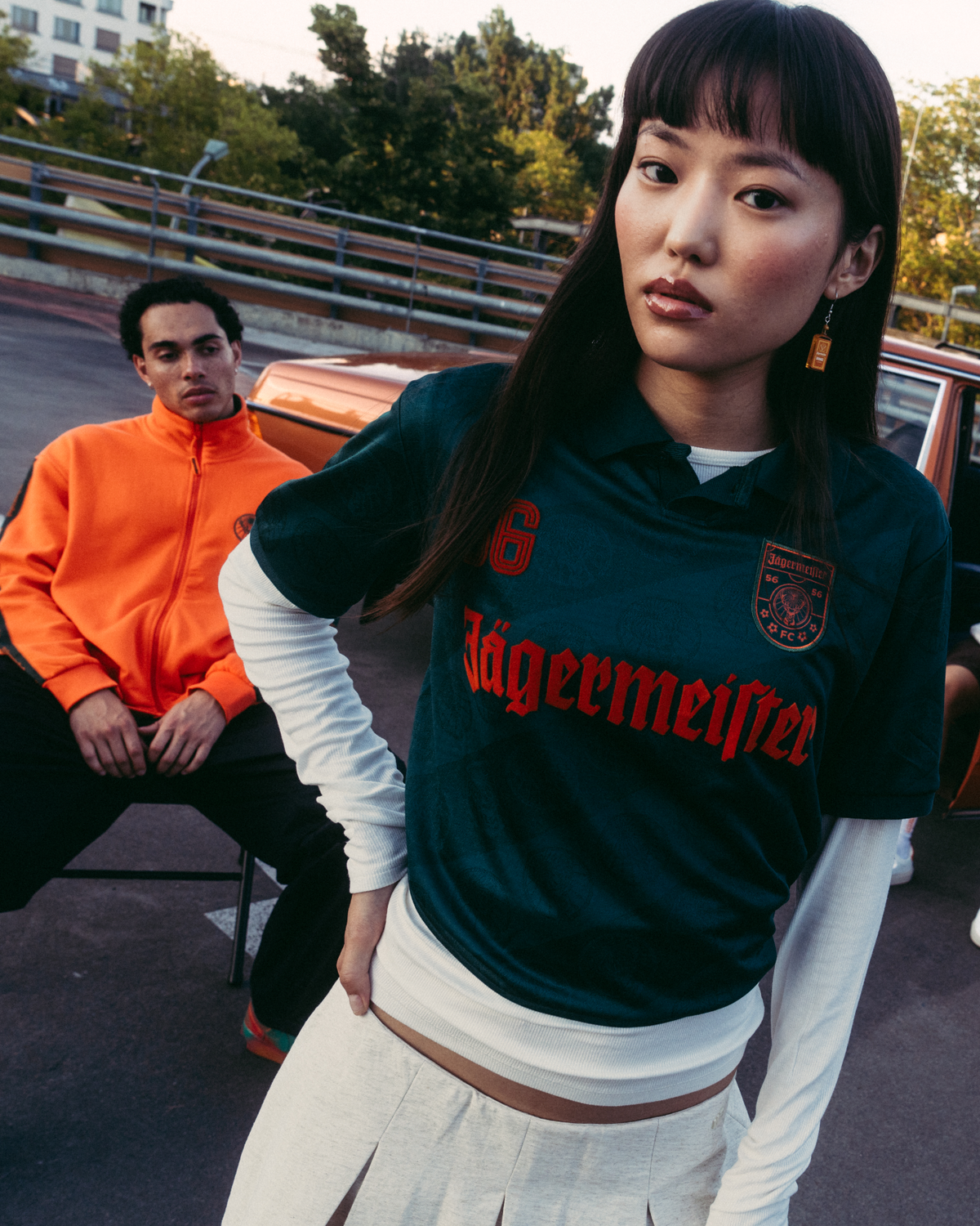 A group of four people posing outdoors in front of a vintage brown car. The central figure is wearing a dark green Jägermeister jersey, while others are dressed in casual streetwear.