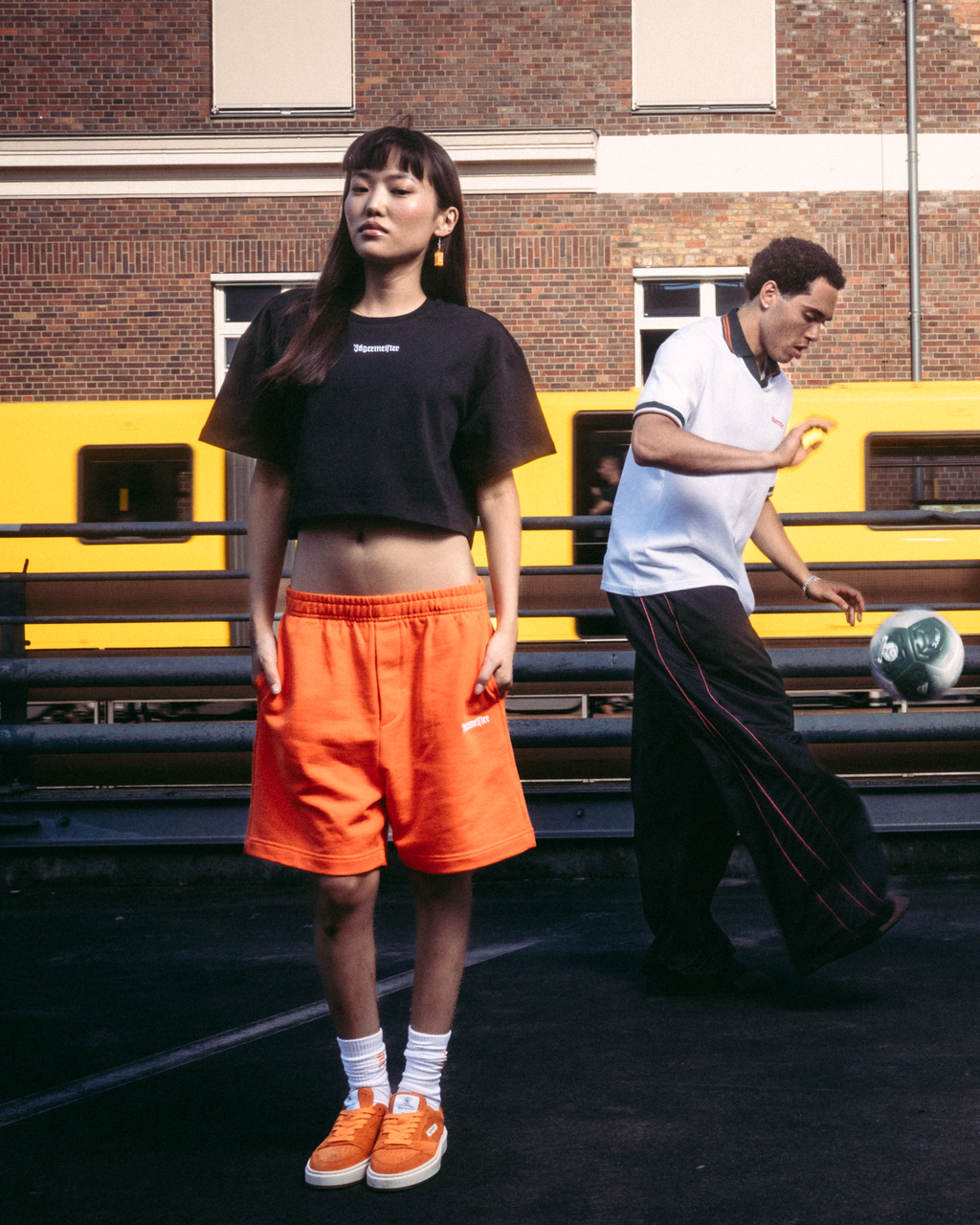 Four models wearing Jägermeister streetwear apparel, including orange shorts, a black crop top, a white t-shirt, and a black hoodie with a graphic back print, standing on a dark paved surface in front of a brick building and a yellow train.