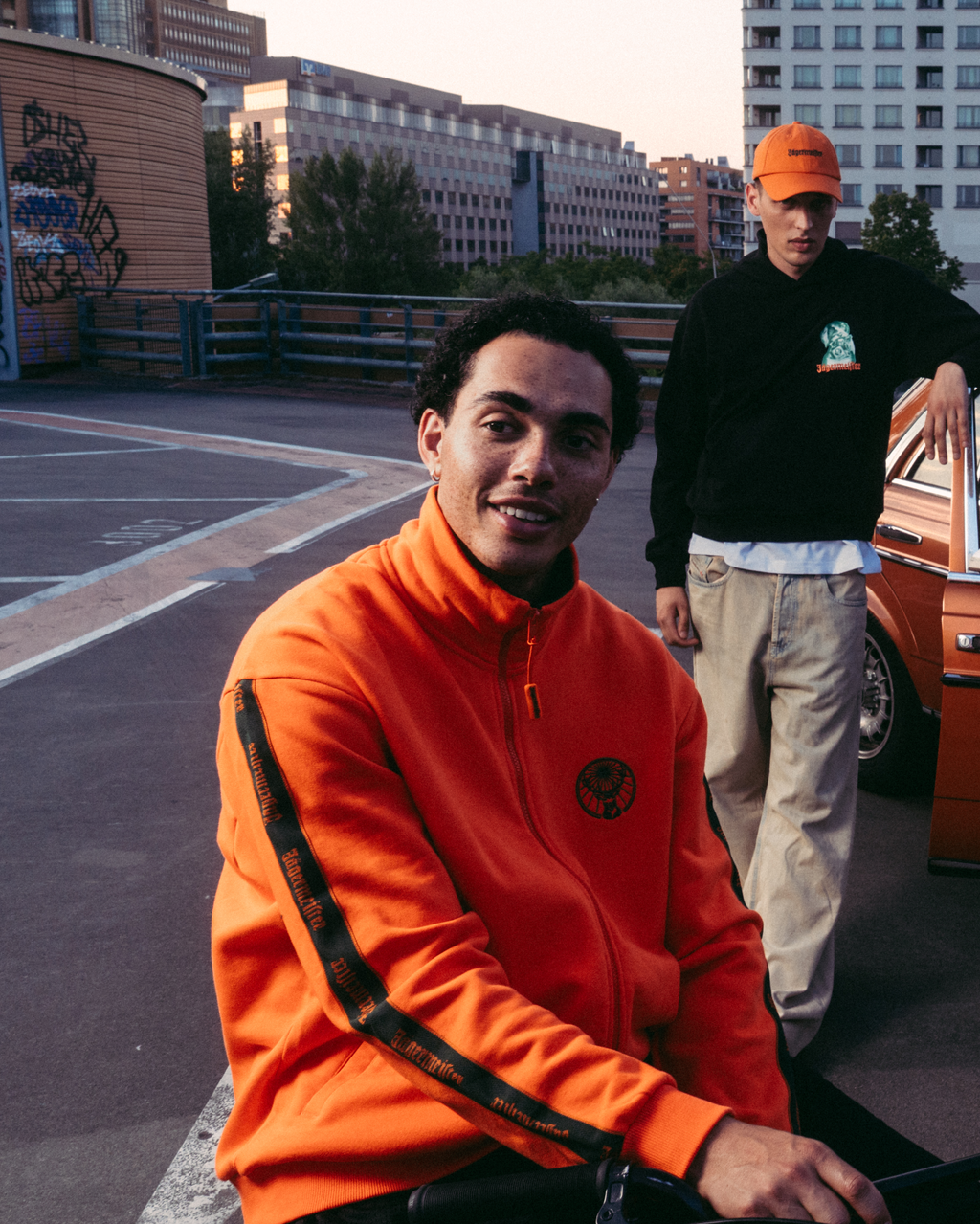 Four models posing in an urban rooftop setting with a vintage orange car, wearing various Jägermeister streetwear outfits including a bright orange Jägermeister zipper, a white Jägermeister graphic t-shirt with Waldi, a dark green Jägermeister jersey, and a white pleated skirt.