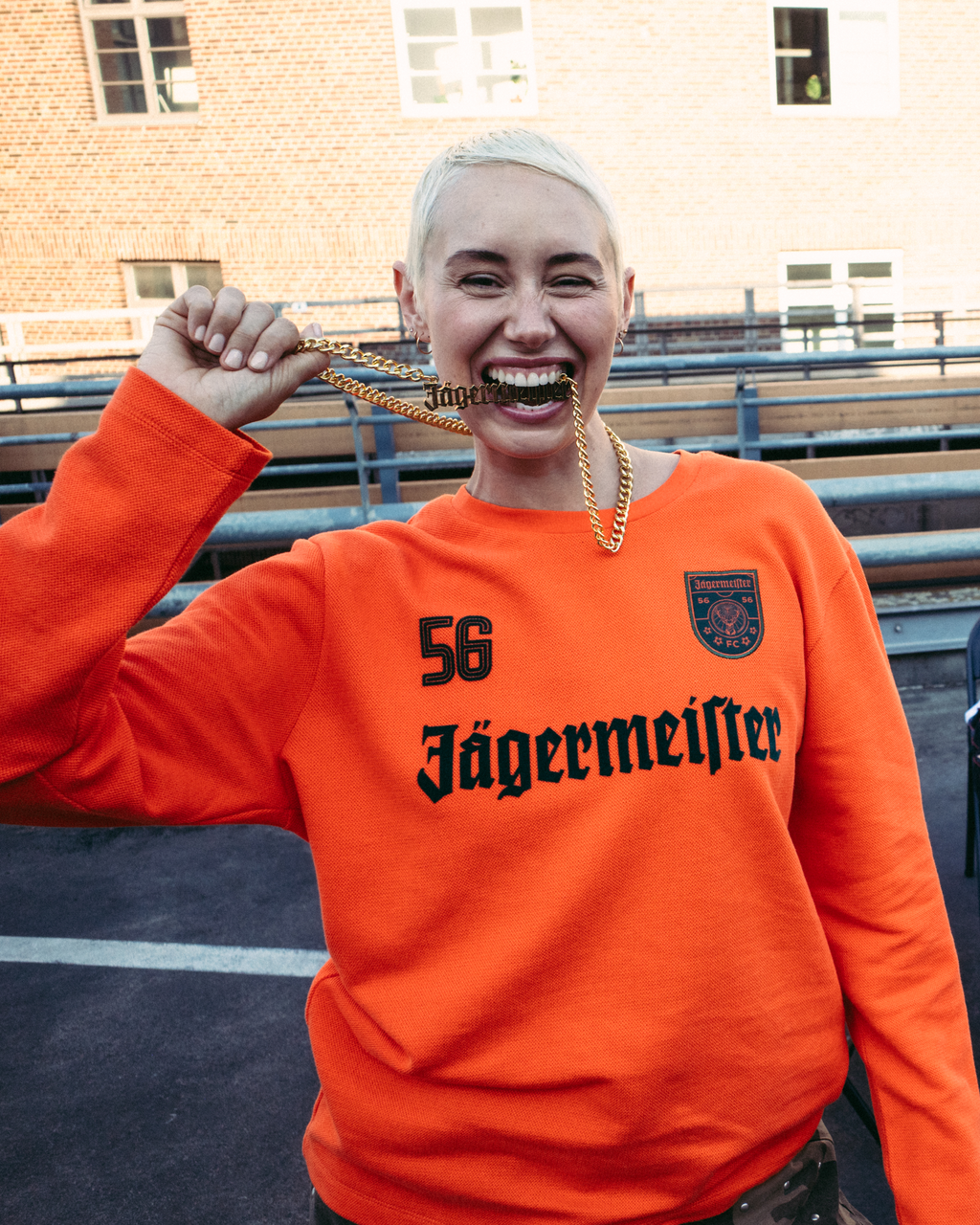A smiling woman in the foreground wears a bright orange Jägermeister long-sleeve sweatshirt with '56 Jägermeister' printed in black on the front, holding a Jägermeister gold chain. 