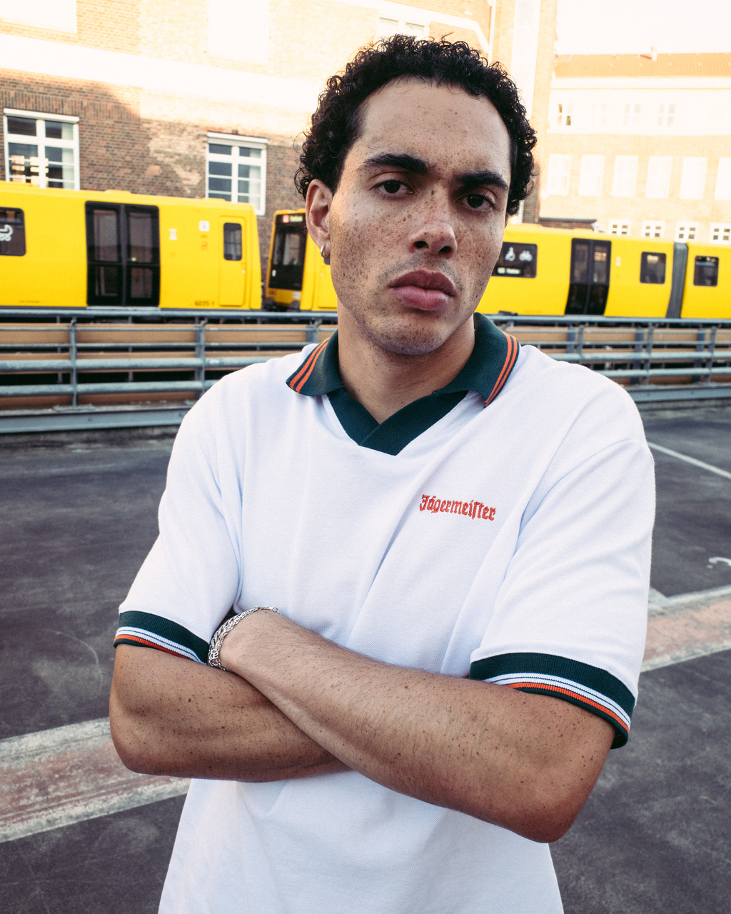 Un homme portant un polo Jägermeister blanc à manches courtes avec liserés vert foncé et orange, avec le nom de la marque « Jägermeister » sur la poitrine, se tient les bras croisés devant des trains jaunes et un bâtiment en brique.