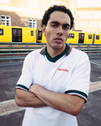 Un homme portant un polo Jägermeister blanc à manches courtes avec liserés vert foncé et orange, avec le nom de la marque « Jägermeister » sur la poitrine, se tient les bras croisés devant des trains jaunes et un bâtiment en brique.