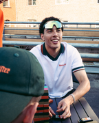 Un homme en polo Jägermeister blanc et vert sourit assis dehors à une table, avec une autre personne portant une casquette verte Jägermeier au premier plan et un bâtiment en brique en arrière-plan.