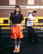 Quatre mannequins portant des vêtements streetwear Jägermeister, notamment un short orange, un crop top noir, un t-shirt blanc et un hoodie noir avec un imprimé graphique dans le dos, debout sur une surface pavée sombre devant un bâtiment en briques et un train jaune.