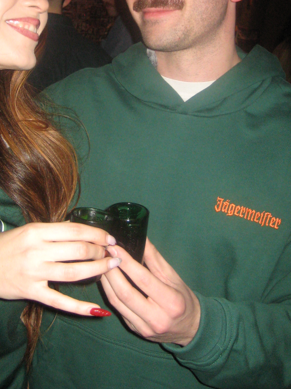 Personne portant un sweat à capuche vert Jägermeister avec le wordmark orange Jägermeister sur la poitrine.
