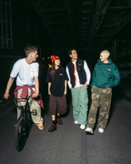 Quatre personnes marchent et roulent à vélo dans une rue sombre la nuit, sous une voie ferrée surélevée, portant des vêtements streetwear Jägermeister de différentes couleurs.