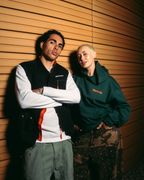 Deux modèles posent devant un mur chaleureux habillé de panneaux de bois. Le modèle de gauche porte un gilet noir Jägermeister par-dessus une chemise blanche à manches longues avec des accents orange, et un pantalon cargo olive.