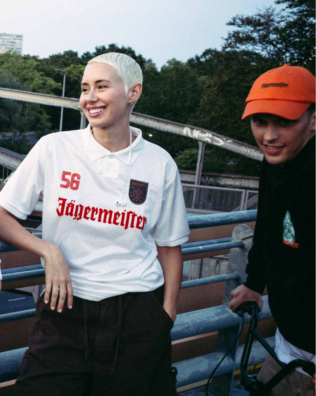 Quatre personnes debout en extérieur sur un pont ou une passerelle, portant des vêtements streetwear Jägermeister. L’une porte une veste zippée orange vif, une autre un maillot vert foncé Jagermeister avec une jupe blanche, une troisième un maillot blanc Jagermeister avec un short foncé, et une quatrième un hoodie noir Jägermeister et une casquette orange.