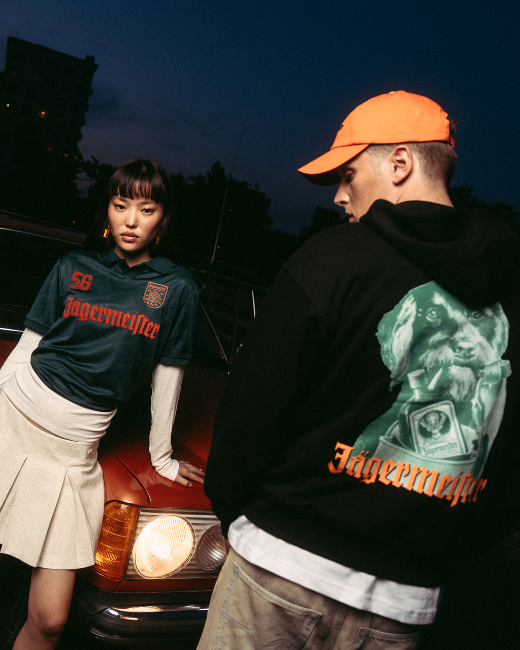 Deux mannequins posent devant une voiture rouge vintage au crépuscule. L’un porte un maillot vert foncé 'Jägermeister' avec une jupe plissée blanche, et l’autre porte un hoodie noir Jägermeister Waldi et une casquette orange.