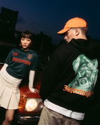 Deux mannequins posent devant une voiture rouge vintage au crépuscule. L’un porte un maillot vert foncé 'Jägermeister' avec une jupe plissée blanche, et l’autre porte un hoodie noir Jägermeister Waldi et une casquette orange.