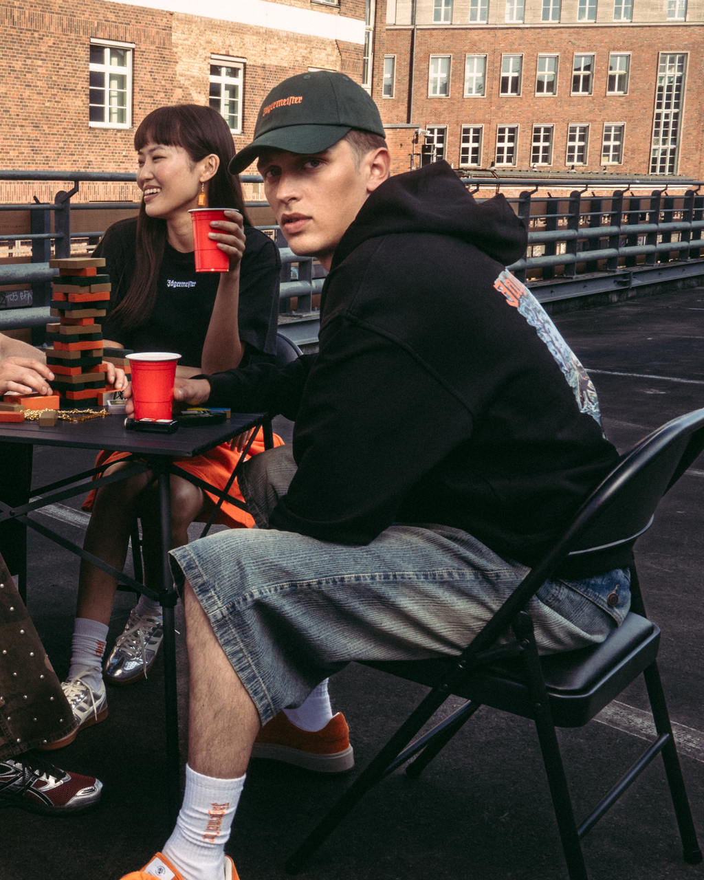 Quatre personnes assises autour d’une petite table sur un toit-terrasse ou un pont en plein air, tenant des gobelets rouges. Elles portent des tenues streetwear Jägermeister décontractées, dont un sweatshirt orange, un t-shirt blanc, un hoodie noir et un pantalon à motif.