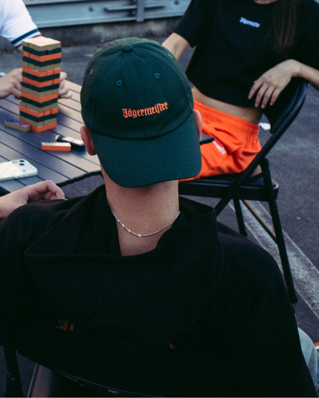Au premier plan, une femme souriante porte un sweatshirt orange vif à manches longues avec « 56 Jägermeister » imprimé en noir sur le devant, et tient une chaîne dorée. Trois personnes sont assises à une table en arrière-plan sur un toit-terrasse ou une plateforme extérieure, avec un immeuble en briques derrière elles.