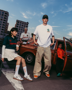 Three models pose with a vintage orange-brown car on an urban street under a blue sky. One model sits on the left in a dark green sweatshirt and white skirt, one stands in the center wearing a white t-shirt and beige pants, and one sits inside the open car door wearing an orange jacket.
