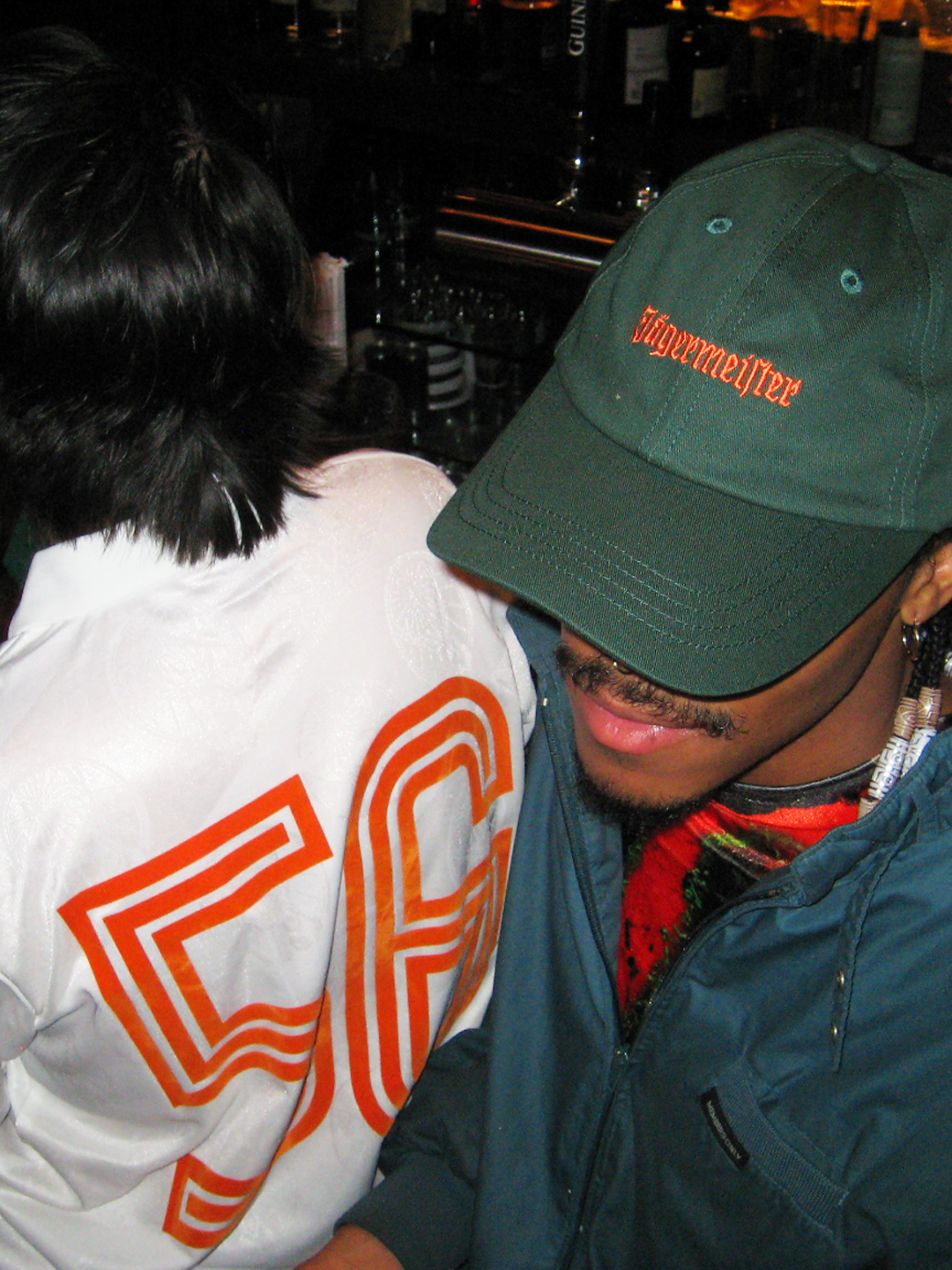 Two people sitting together in a bar setting, one wearing a green Jägermeister dad cap.