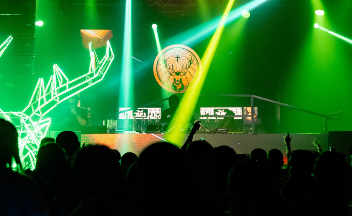 Crowd gathered in front of a DJ booth at a live event, illuminated by green stage lighting and laser beams, with the Jägermeister stag logo displayed prominently in the background.