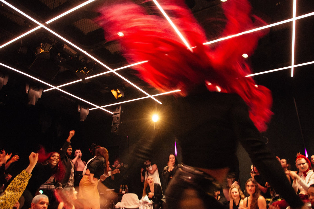 A person with very bright red hair wearing a black outfit, captured mid-motion in a crowded indoor venue with glowing overhead linear lights and a crowd below.