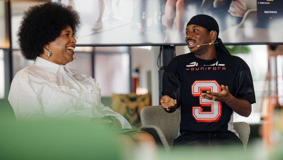 A man wearing a black youth jersey with a large red and white number 3 and the brand name 'YOUNIFORM' sits in a chair, smiling and gesturing while talking to a woman in a white blouse.