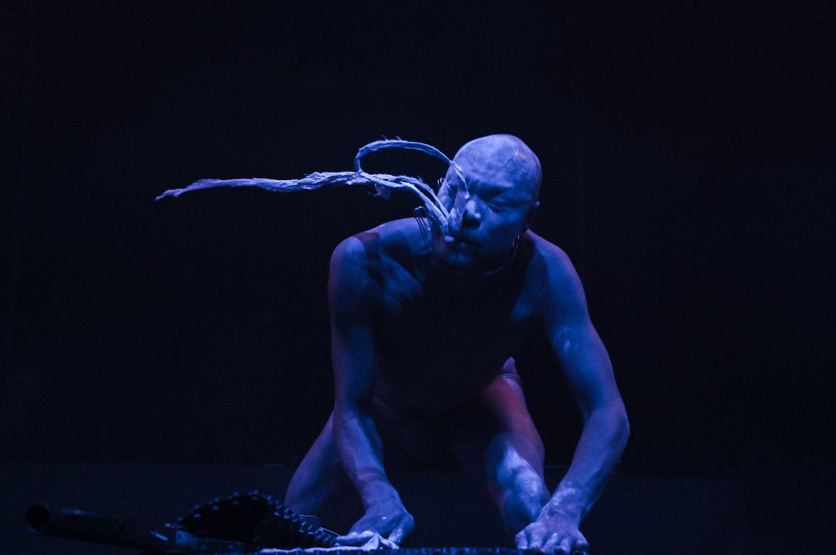 A dancer or performer crouches on a dark stage under dramatic blue lighting, with a long branch-like prop extending from the mouth area and hands reaching forward.