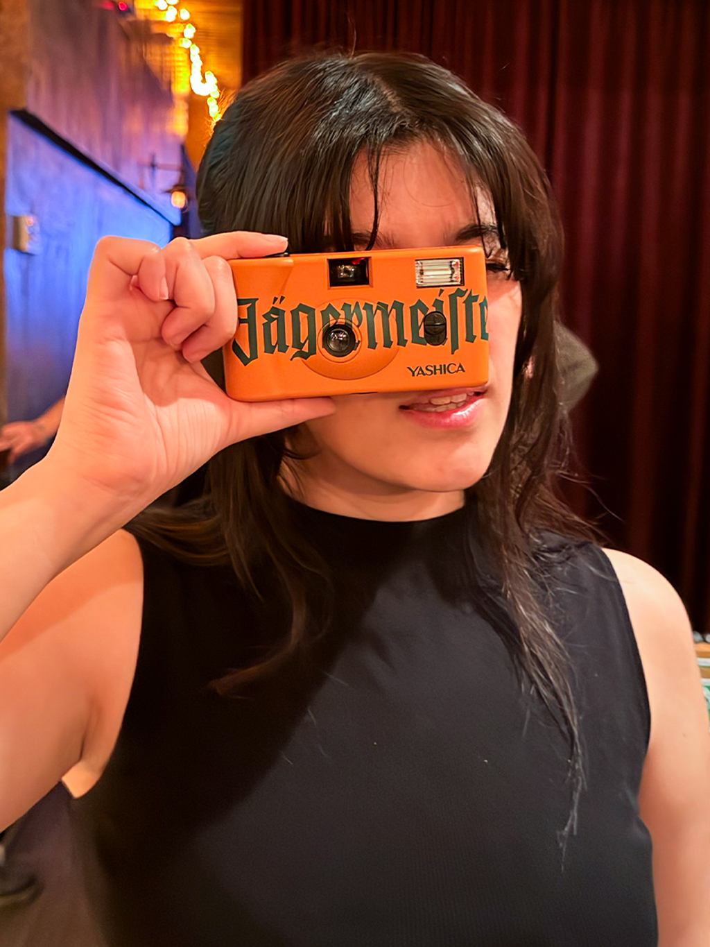 Personne tenant un appareil photo orange avec le marquage « Jägermeister » devant son visage.
