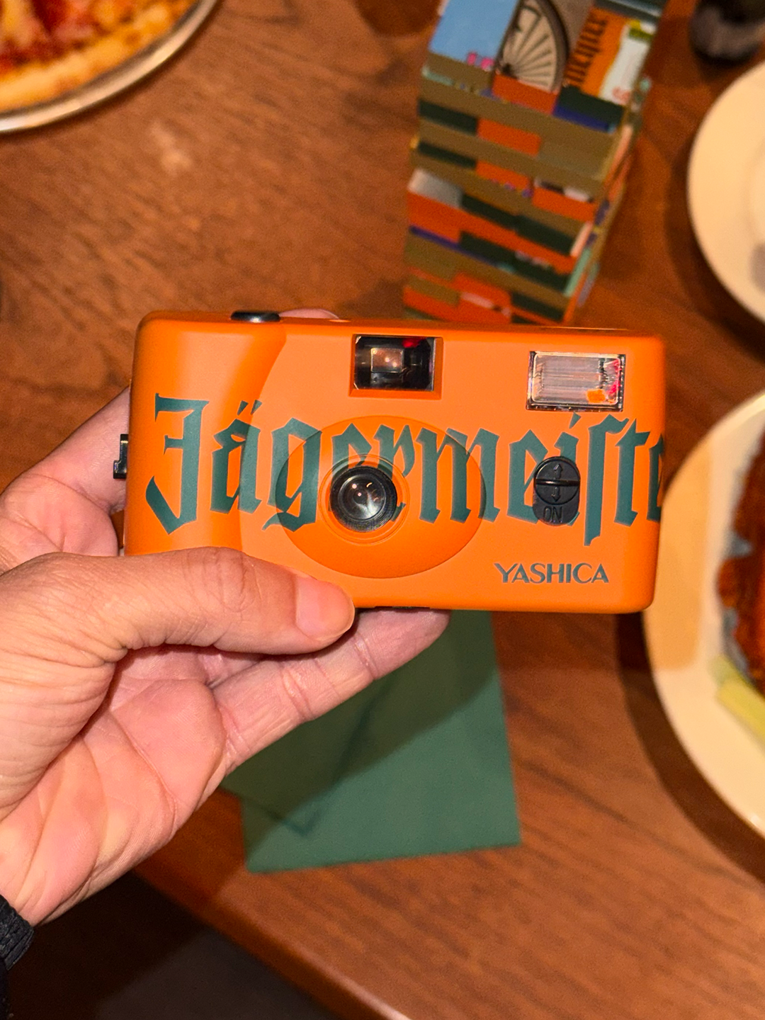 Appareil photo orange avec le marquage « Jägermeister » tenu par une main sur une table en bois.