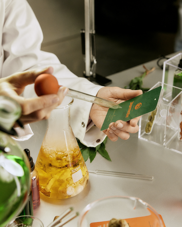 Une personne tenant une bouteille de liquide jaune avec une carte parfumée verte, entourée d’objets de laboratoire.