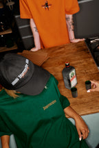 Personne portant une chemise verte « Jägermeister » et une casquette noire Jägermeister Racing Cap, assise à une table sur laquelle se trouvent des produits Jägermeister.