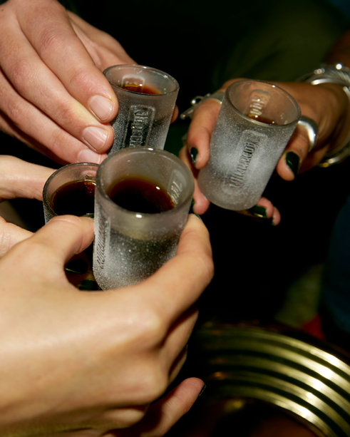 Quatre mains tenant des verres à shot Jägermeister givrés remplis d’un liquide foncé, en train de trinquer.