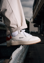 A person wearing off-white Jägermeister sneakers with white socks and beige pants, sitting on a metal railing in an outdoor urban setting.