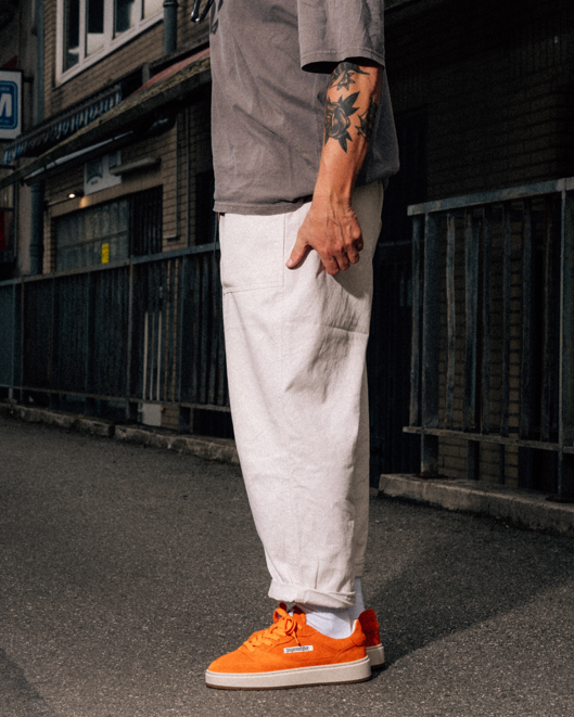 A person standing outdoors on a street wearing loose white trousers, a gray t-shirt, and bright orange low-top sneakers. The photo is cropped from the chest down, with the person's right arm visible and the background showing a dark urban alley or street setting.